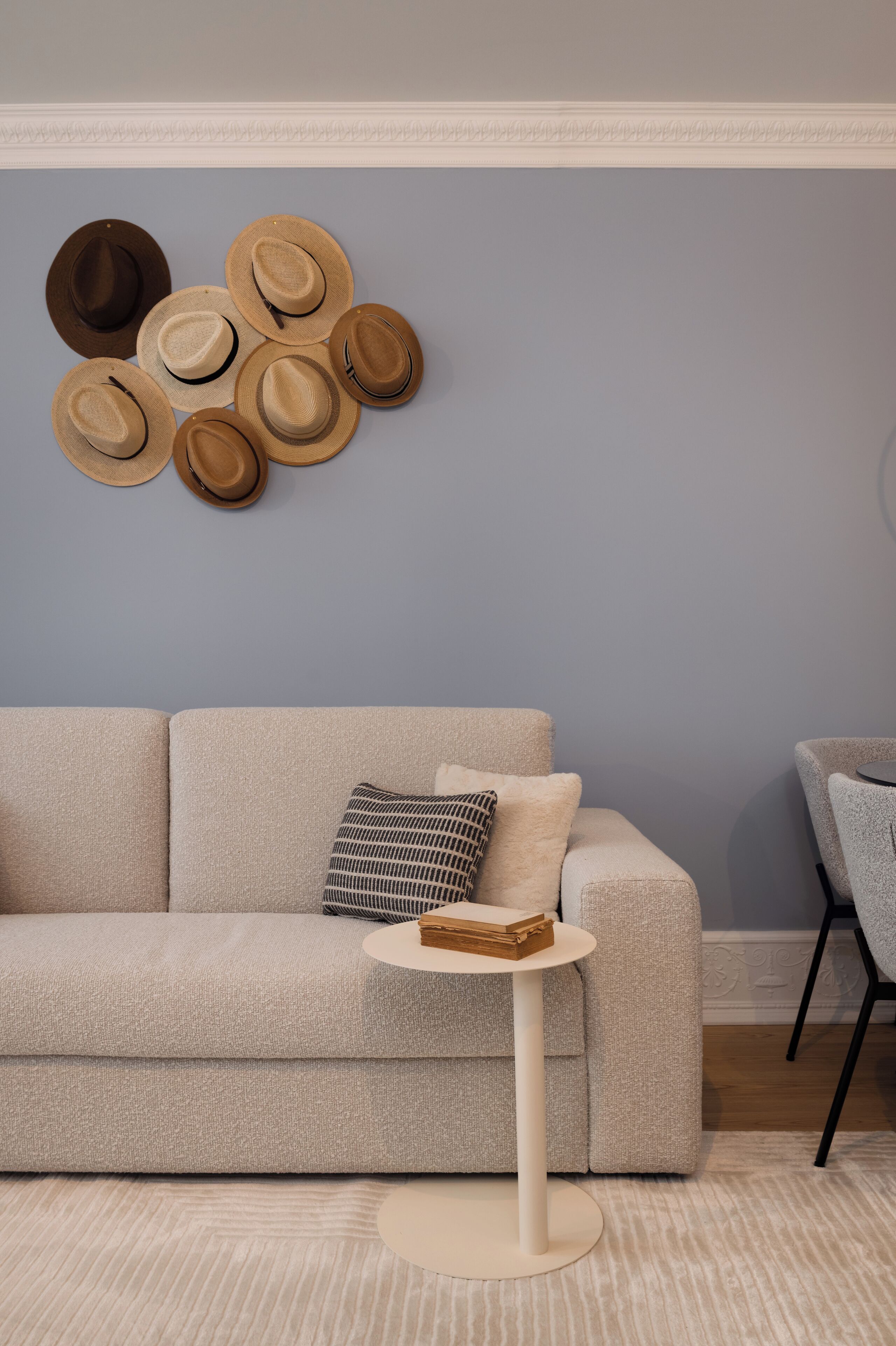 Un sofá color beige con un cojín estampado, una mesita redonda con una cesta, y una pared decorada con sombreros.