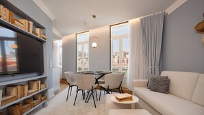 A modern living room with a flat-screen TV, bookshelves, a sofa, a dining table with chairs, and large windows with curtains.