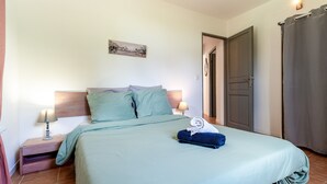 Un lit bien fait, recouvert d’une couverture bleue, une table de chevet en bois avec une lampe, et une porte sur la droite.