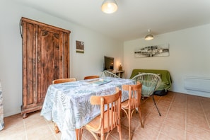 Ein Essbereich mit einem Tisch für vier Personen, Holzstühlen, einem Schrank, einem Bett mit grüner Bettdecke und einem an der Wand hängenden Bild.