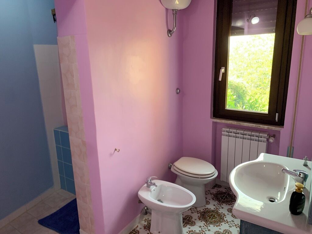 A bathroom with a pink wall, a white sink, a bidet, a toilet, and a window with a view of greenery.