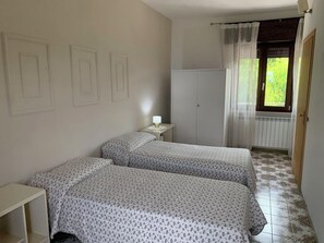 A bedroom with two beds, a wardrobe, a bedside table with a lamp, and a window with curtains. - Small villa with a view of the Cassino valley and montecassino with its abbey. (San Bartolomeo)
