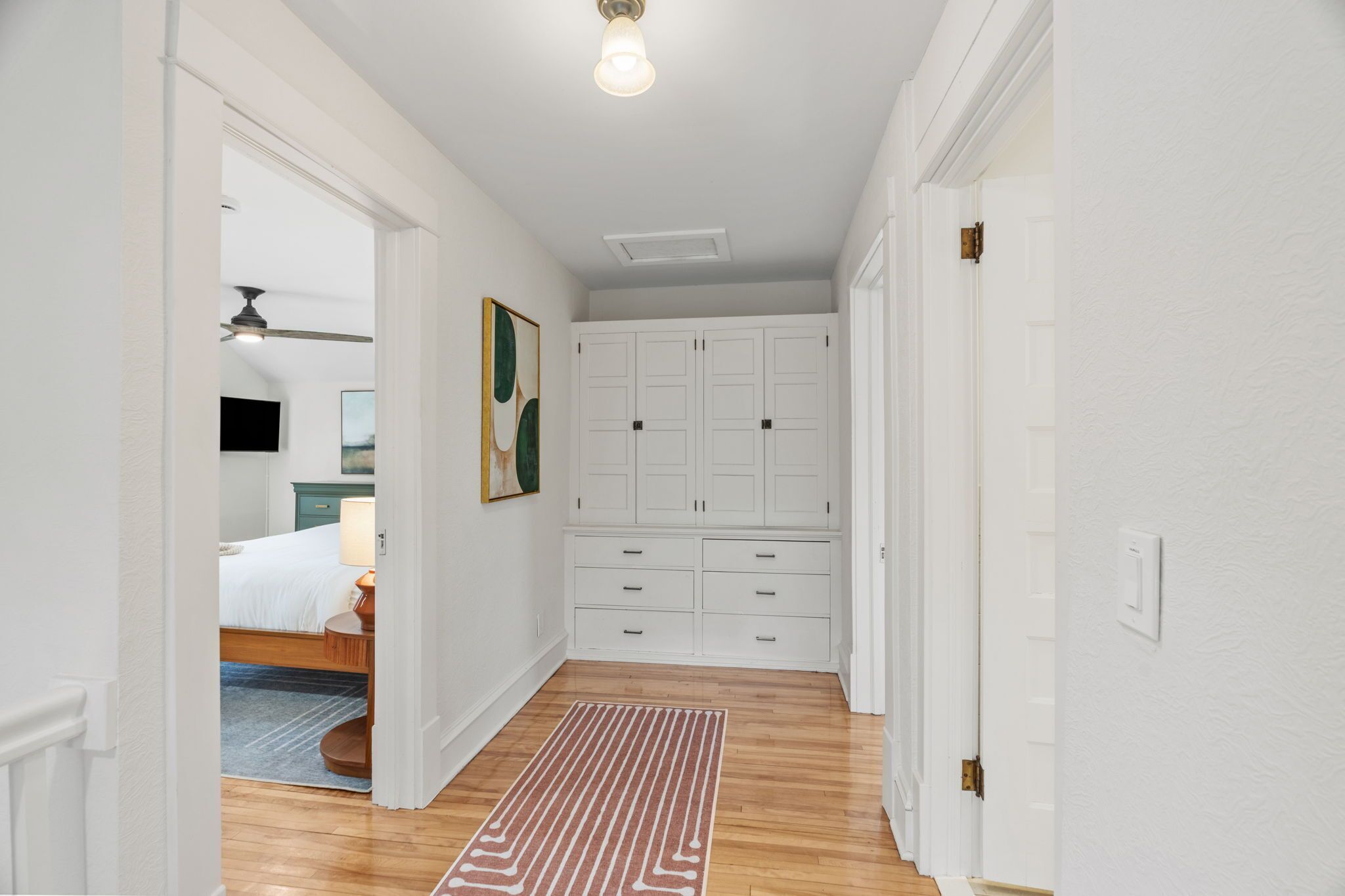 Een gang met houten vloer, witte muren, een plafondventilator en een deur die leidt naar een slaapkamer met een bed en nachtkastje.