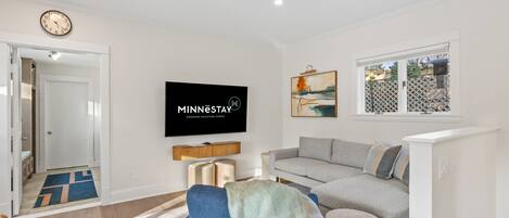 A modern living room with a flat-screen TV, a gray sofa, a blue armchair, and a window with a view.