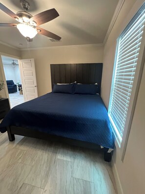 Un dormitorio con colcha azul oscuro, ventilador de techo, ventana con persianas y puerta que da a otra habitación.