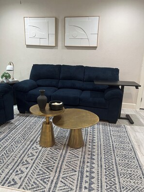 Una sala de estar moderna con un sofá azul oscuro, una mesa de centro redonda de latón y una alfombra estampada. Hay dos obras de arte abstracto en la pared sobre el sofá.