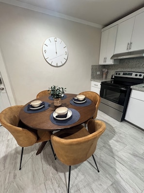 Un comedor con una mesa redonda dispuesta para cuatro personas, rodeada por cuatro sillas. Un reloj está colgado en la pared sobre la mesa.