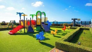 A playground with slides and climbing structures, a well-maintained lawn, and a clear view of the sea in the background. - Magnificent sea-view and waterfront apartment at Tamaris Dar Bouazzza (Tamaris)