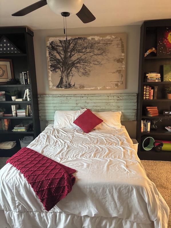 A bedroom with a bed, a ceiling fan, a bookshelf, and a wall-mounted painting.