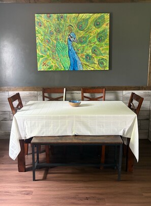 A dining table with a white tablecloth, wooden chairs, and a peacock painting on the wall. - Sage & Sky - Ranch Style Home (Hays)