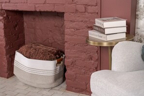 A room with a brick fireplace, a grey armchair, a small round table with books, and a white basket with a brown blanket. - Boutique Italian Village OSU 4BR + 4 parking spots (Columbus)
