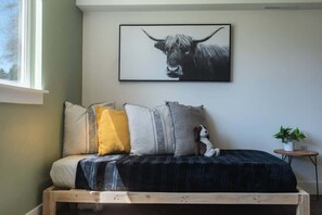 Un lit recouvert d’une couverture sombre, plusieurs oreillers, un petit chien, un tableau encadré représentant un taureau et une petite table sur laquelle se trouve une plante.