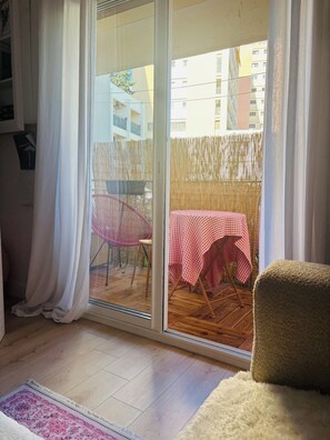 Una habitación con puerta corrediza de vidrio que da a un balcón con mesa y sillas, piso de madera y una alfombra blanca y mullida.
