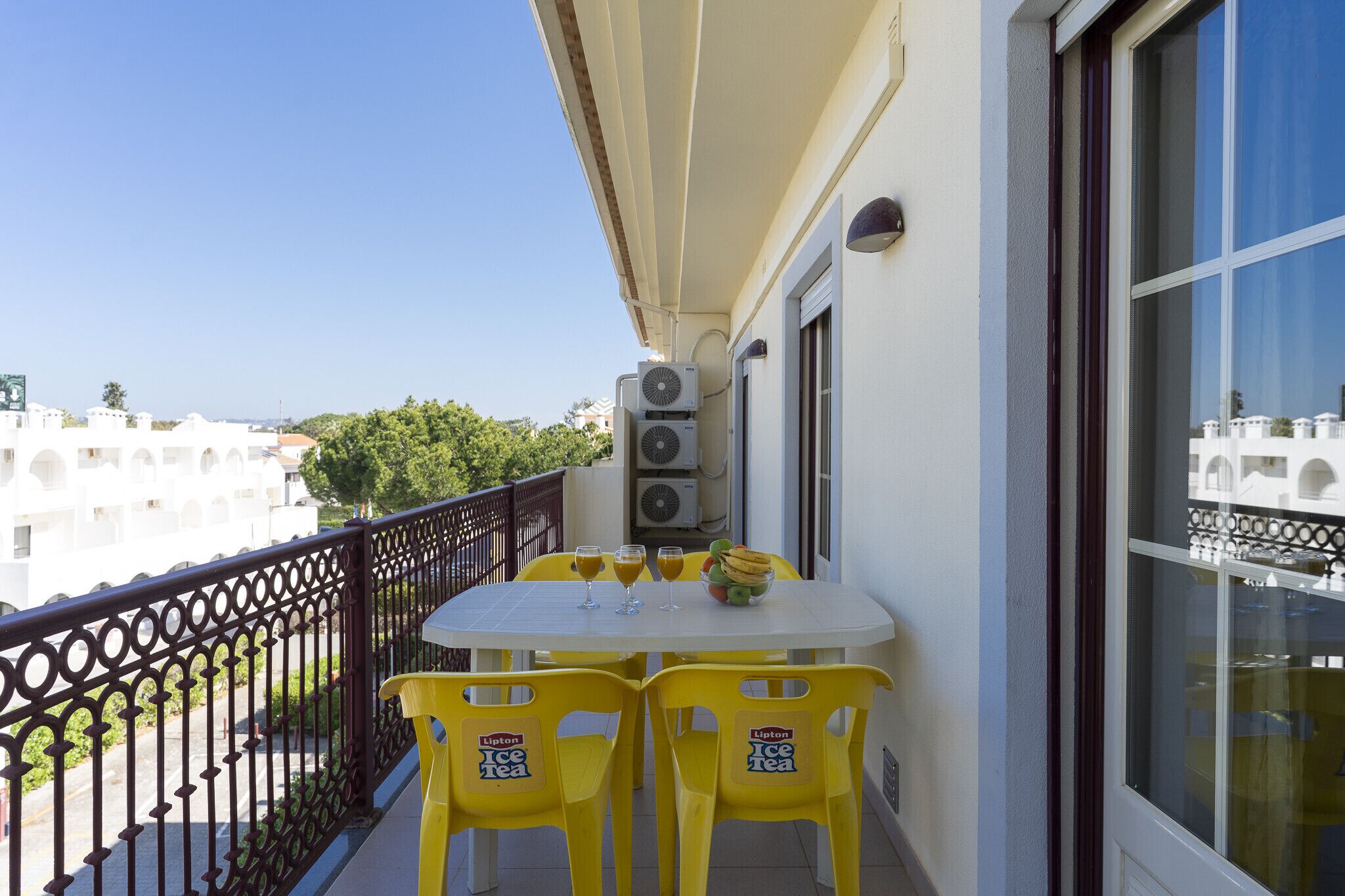 Un balcón con mesa y sillas, unidades de aire acondicionado y vistas a edificios y árboles.