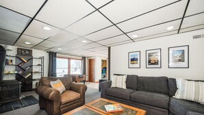 En stue med en læderlænestol i brunt, en grå sofa og et træbord. Væggen er pyntet med billeder i rammer, og et reol står med bøger og pynteting.