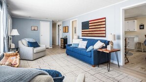 En stue med en blå sofa, en grå lænestol og et sofabord. Der hænger et flag på væggen, og en dør fører til et andet rum.