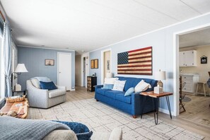 Un salon avec un canapé bleu, un fauteuil gris et une table basse. Un drapeau est accroché au mur, et une porte donne accès à une autre pièce.