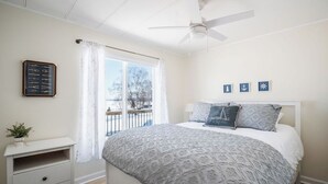 Et soveværelse med seng, natbord, ventilator i loftet, et vindue med gardiner og udsigt til det fri.