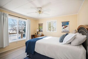 Une chambre à coucher avec un lit, un ventilateur de plafond, une fenêtre avec des rideaux et un mur orné d’un tableau encadré.