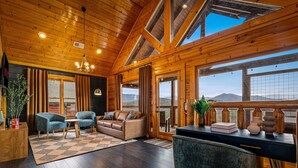 Una habitación acogedora con paredes de madera, un sofá, dos sillones, un escritorio y ventanas amplias con vistas a la montaña.
