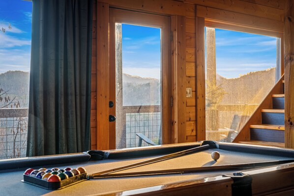 Une table de billard avec des billes et une queue, une fenêtre avec des rideaux et une vue sur un paysage montagneux.