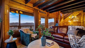 Interior acogedor de una cabaña de madera con un sofá de cuero, una mesa redonda de centro y vistas a las montañas a través de amplios ventanales.