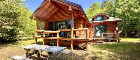 A rustic log cabin with a porch and picnic tables, surrounded by trees.