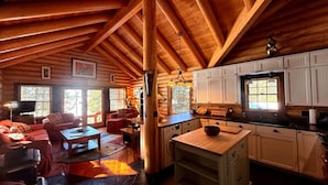 Un intérieur de chalet chaleureux comprenant un salon, un coin repas et une cuisine, avec des poutres en bois et de grandes fenêtres.