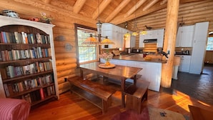 L’intérieur chaleureux d’un chalet en bois comprend une bibliothèque en bois, une table à manger avec des bancs, un coin cuisine et une grande fenêtre qui laisse entrer la lumière naturelle.