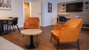 A modern living room with a round wooden table, two orange armchairs, a black floor lamp, and a TV mounted on the wall. - Hotel & Restaurant Wefers (Emsdetten)