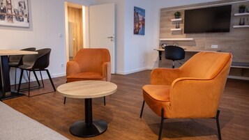 Una sala de estar moderna con una mesa redonda de madera, dos sillones naranja, una lámpara de pie negra y un televisor montado en la pared.