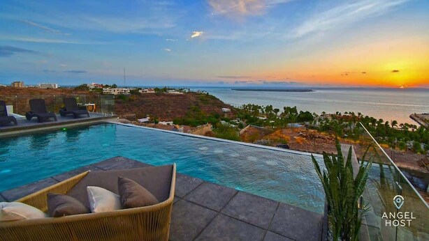 Ein Poolbereich mit Blick auf den Sonnenuntergang über dem Meer, Liegestühle und eine entfernte Küstenlinie.