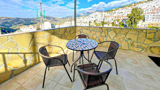 Ein Balkon mit einer Steinmauer, einem runden Tisch mit Stühlen und Blick auf eine Küstenstadt.