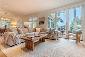 A spacious living room with a large sectional sofa, a wooden coffee table, and a view through large sliding glass doors to an outdoor patio. - Walk to Beach + Golf Views | Palmetto Dunes (Hilton Head Island)