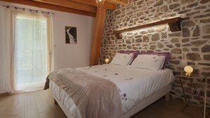 Une chambre avec un mur en pierre, un lit avec une literie blanche et des oreillers violets, un plancher en bois et une fenêtre ornée de rideaux légers.