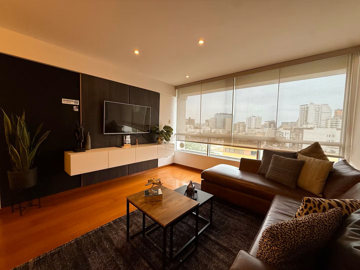 A modern living room with a flat-screen TV, a sofa, a coffee table, and a large window with a city view.