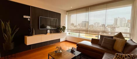 Una sala de estar moderna con una televisión de pantalla plana, un sofá, una mesa de centro y un ventanal con vistas a la ciudad.