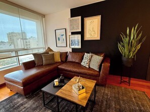 Un salon moderne avec un canapé d’angle en cuir marron, une table basse en bois et une grande fenêtre donnant sur la ville.