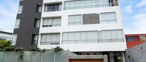 A multi-story building with a modern design, featuring a combination of white and dark grey facades, large windows, and a wooden garage door.