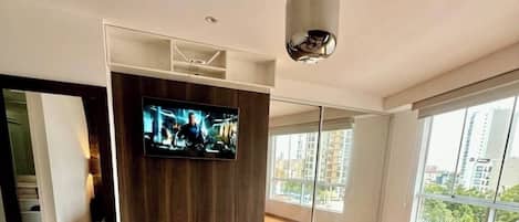 Ein modernes Schlafzimmer mit einem großen Bett, einem an einer Holzvertäfelung montierten Flachbildfernseher und Blick auf die Stadt durch große Fenster.