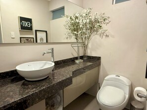 Une salle de bain avec un lavabo blanc, des toilettes et un vase rempli de fleurs blanches posé sur le comptoir.