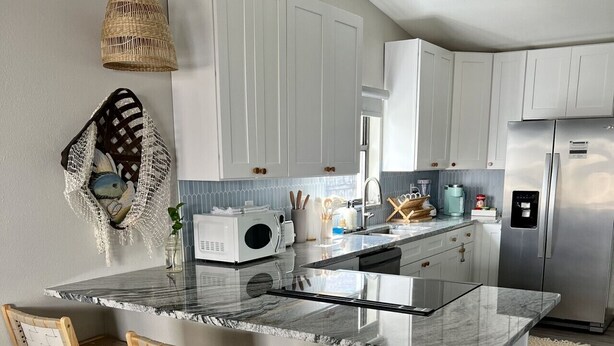 A modern kitchen with white cabinets, a large island with bar stools, a hanging light fixture, and a stainless steel refrigerator.