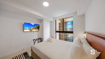 A hotel room with a bed, a television mounted on the wall, a window with a view of a building, and a wooden headboard.