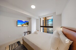 Une chambre d’hôtel avec un lit, un téléviseur fixé au mur, une fenêtre donnant sur un immeuble et une tête de lit en bois.