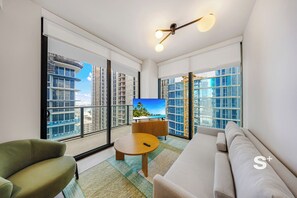 A modern living room with a sofa, armchair, and coffee table, large windows with a city view, and a television on a wooden cabinet. - Modern Condo in DTW Miami - Pool & Gym (Miami)