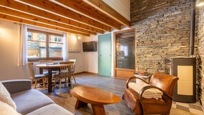 A cozy living room with a wooden ceiling, stone wall, and a dining area with chairs and a table. - L’Écrin des Neiges - Proche Station de Ski (Fontrabiouse)