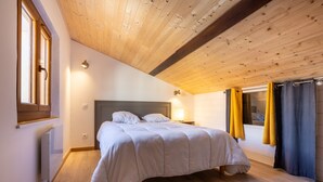 A bedroom with a wooden ceiling, a bed with white bedding, a nightstand with a lamp, and a window with curtains. - L’Écrin des Neiges - Proche Station de Ski (Fontrabiouse)