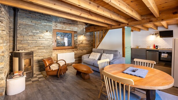A cozy cabin interior with a stone wall, wooden ceiling beams, a stone fireplace, a sofa, a dining table, and a kitchen area. - L’Écrin des Neiges - Proche Station de Ski (Fontrabiouse)