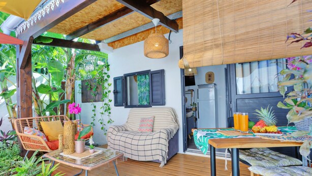 A cozy outdoor seating area with a wooden deck, a wicker chair, a cushioned sofa, and a table with fruits and drinks. - Ti Caz la Zac - Parking Privé (La Riviere)