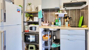 A compact kitchen with a built-in microwave, oven, and a small refrigerator. There are shelves with plants and kitchen utensils, and a sink with a faucet. - Ti Caz la Zac - Parking Privé (La Riviere)